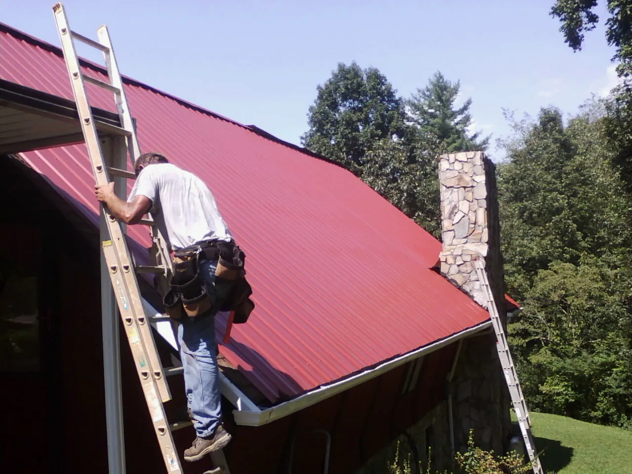 Roofer on a ladder beside a red metal roof and stone chimney — Soffit & Fascia Repair by Roofing Brothers in Swannanoa